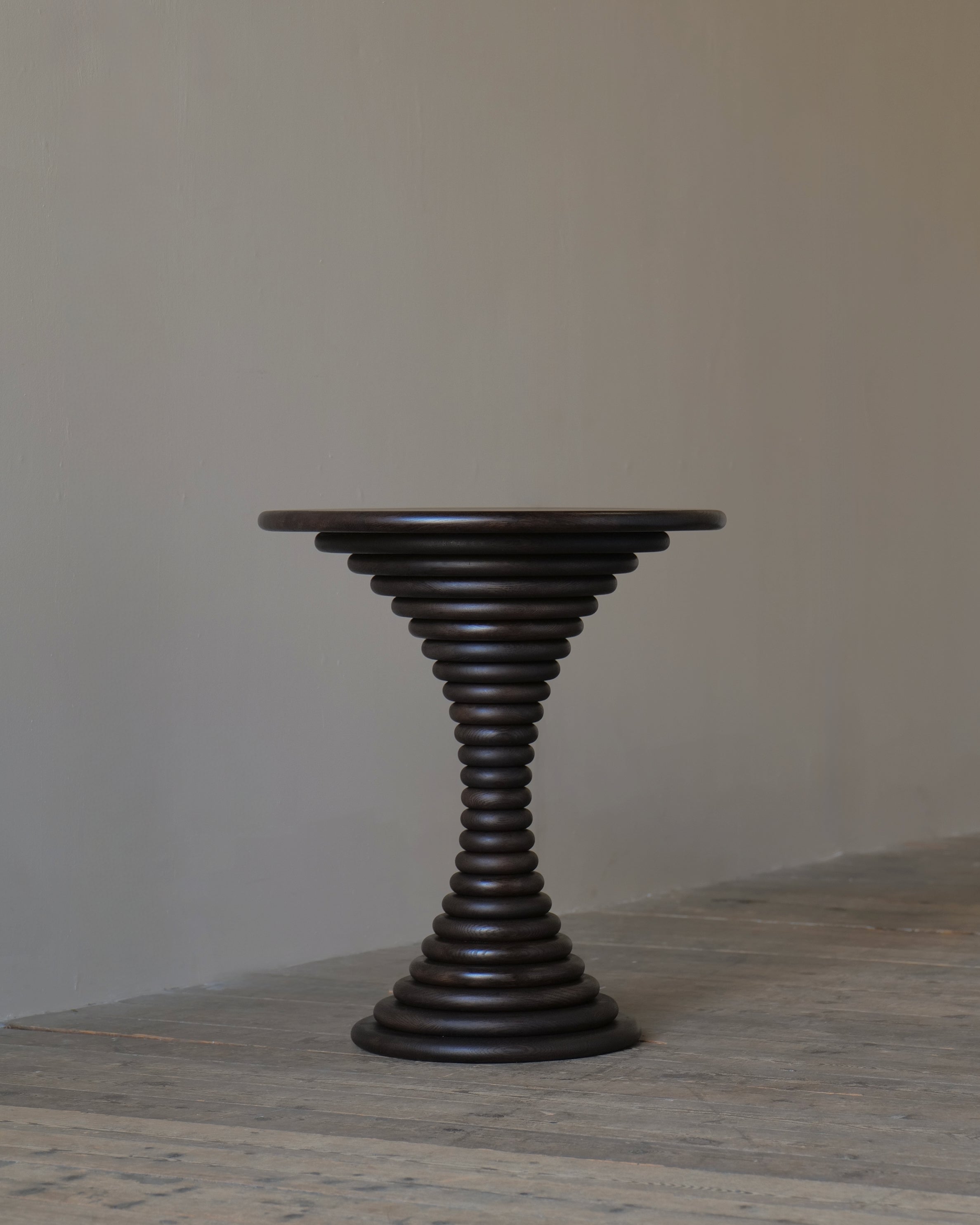 A dark brown wooden pedestal table with a circular top, set against a neutral beige wall and textured grey floor.
