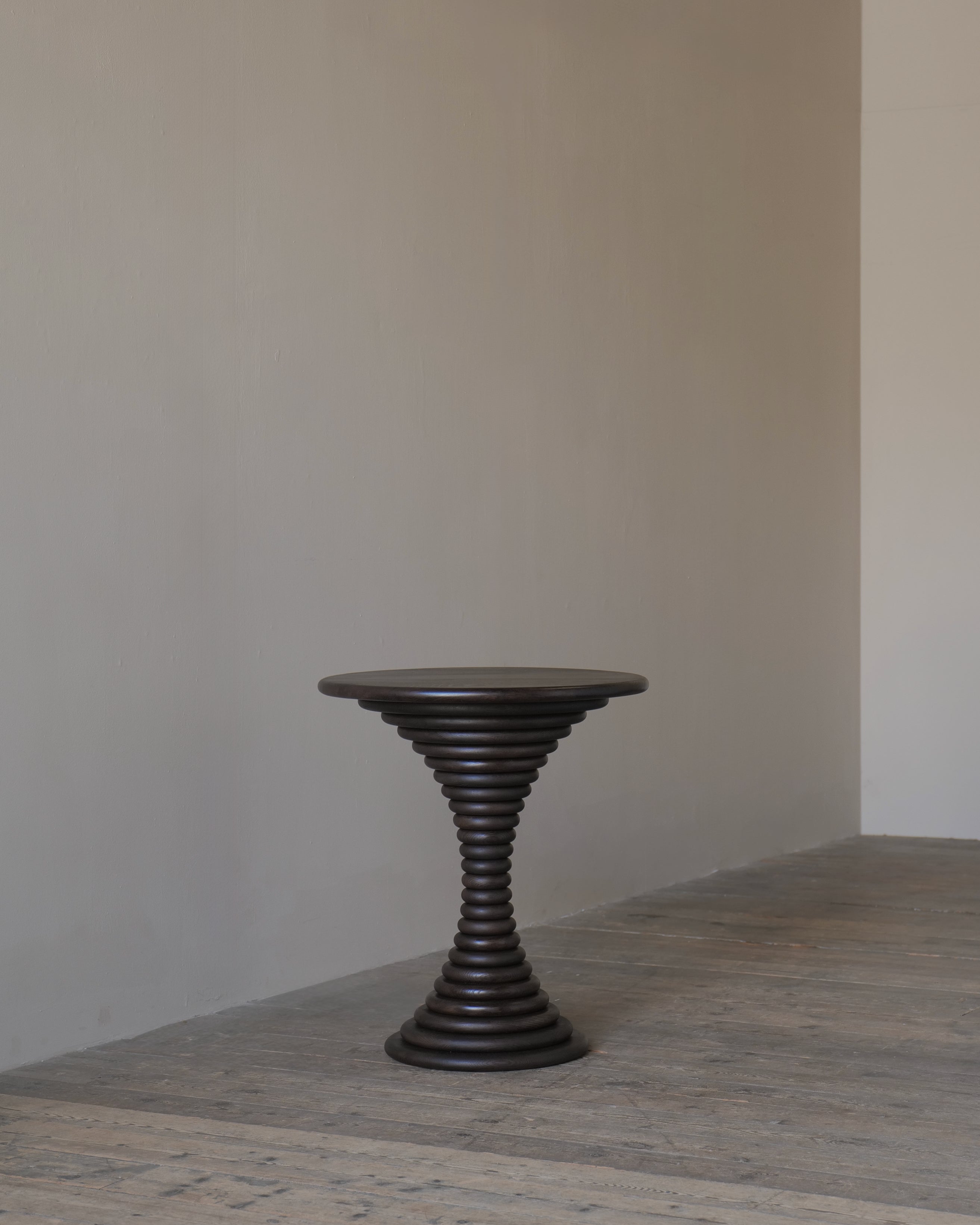 A dark brown timber pedestal table with a circular top, set against a minimalist beige wall and textured grey floor.