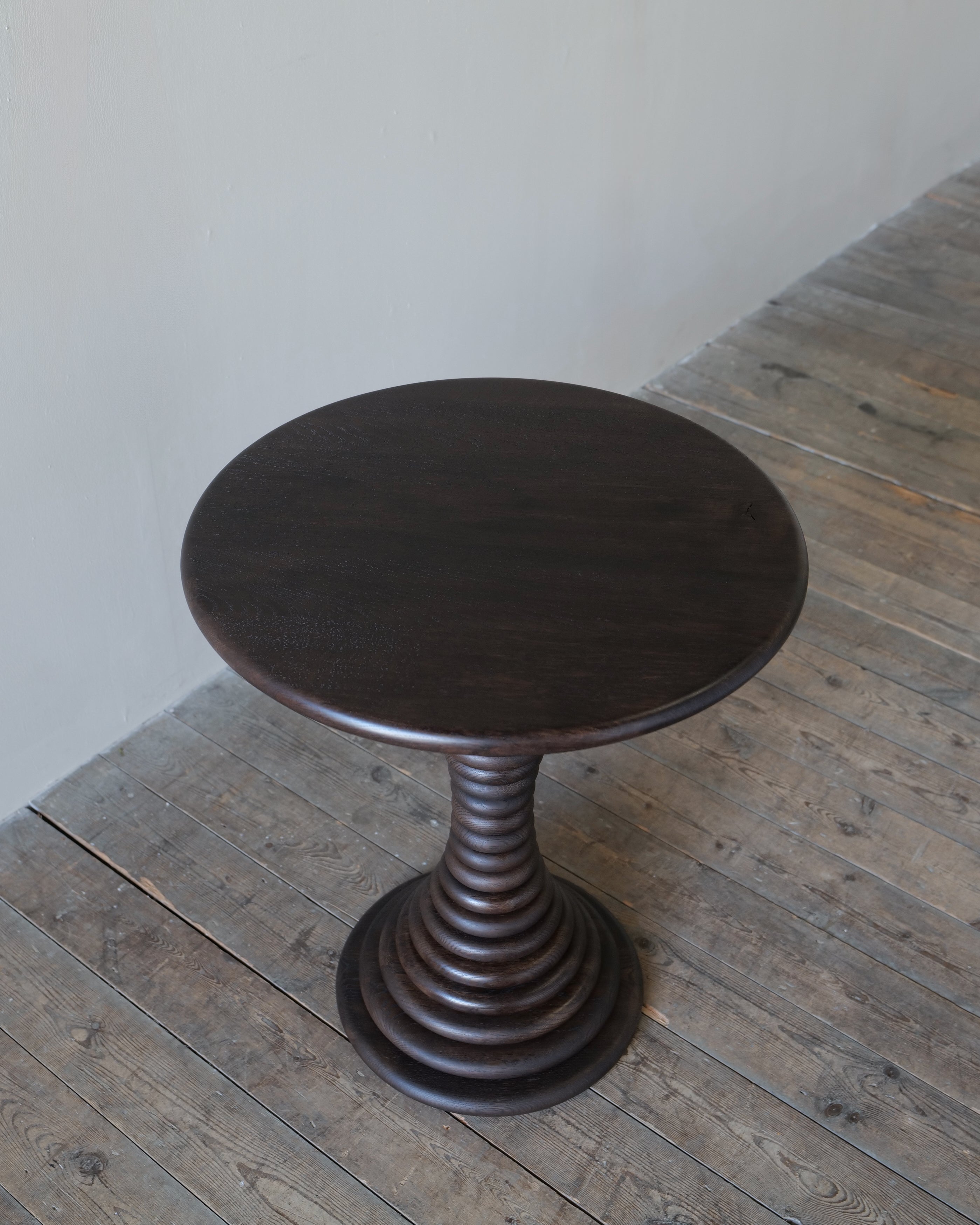 A circular dark brown timber side table with a pedestal base, set on a rustic wooden plank floor against a plain off-white wall.