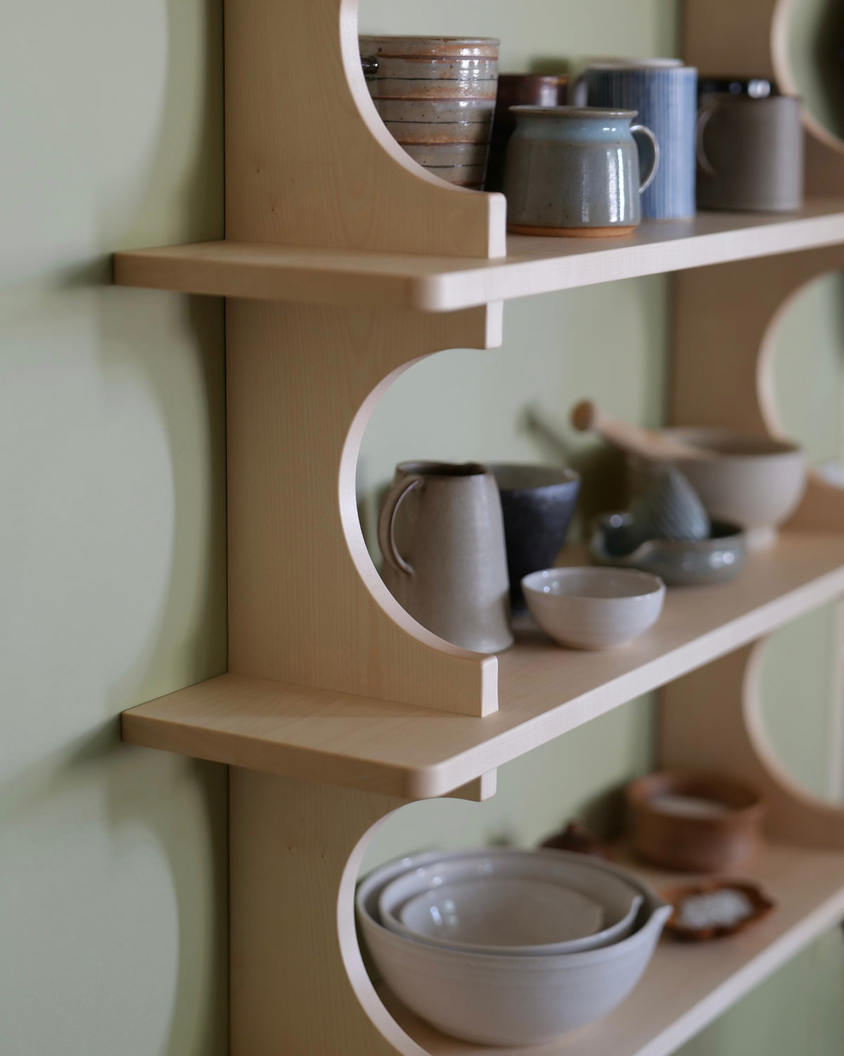 A light natural wood floating shelf with circular cutouts, styled with ceramic mugs, bowls, and cups against a pale green wall.