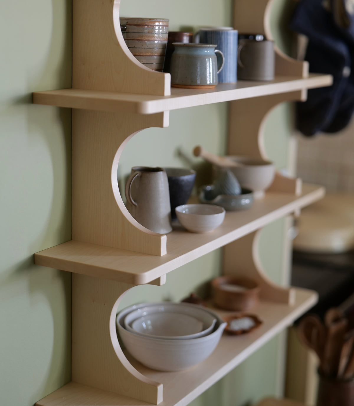 A light natural wood wall-mounted shelving unit with three curved open shelves, styled with ceramic mugs, bowls, and plates against a pale green wall.