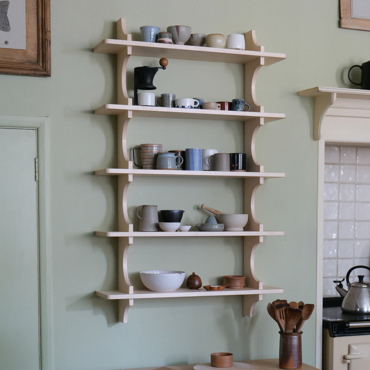 A set of five light natural wood wall shelves with decorative curved supports, mounted on a sage green wall in a kitchen setting. The shelves are styled with various ceramic mugs, bowls, and cups.