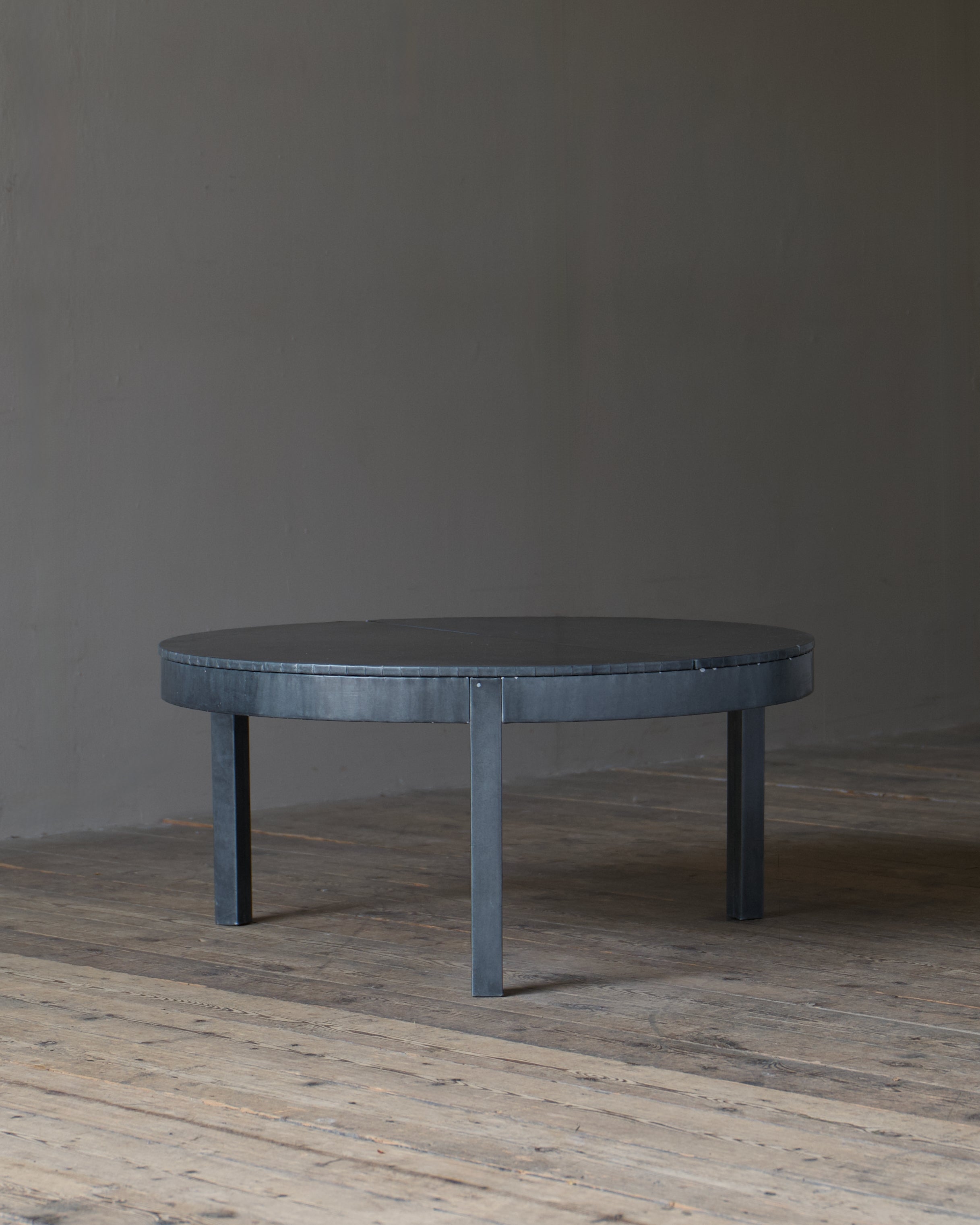 A circular dark zinc wrapped table with four legs, set on a rustic wooden floor against a plain grey wall.