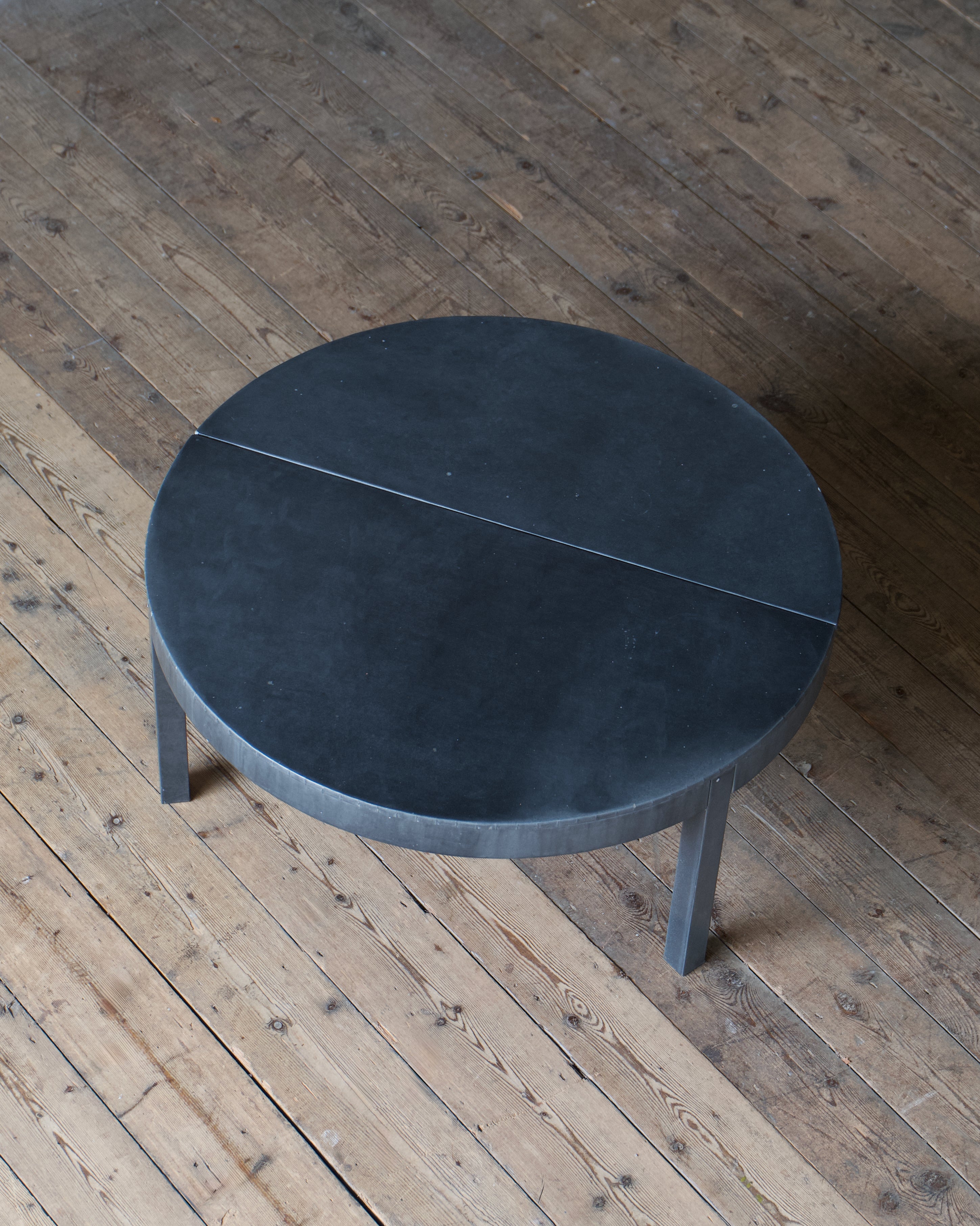 A circular zinc table with a split-top design, photographed from an elevated angle on a rustic wooden plank floor.