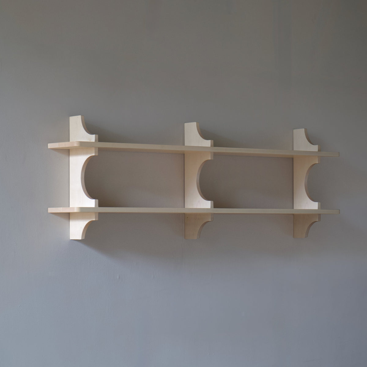 A minimalist three-tier wooden wall shelf with curved style brackets, mounted on a plain grey wall above a wooden floor.