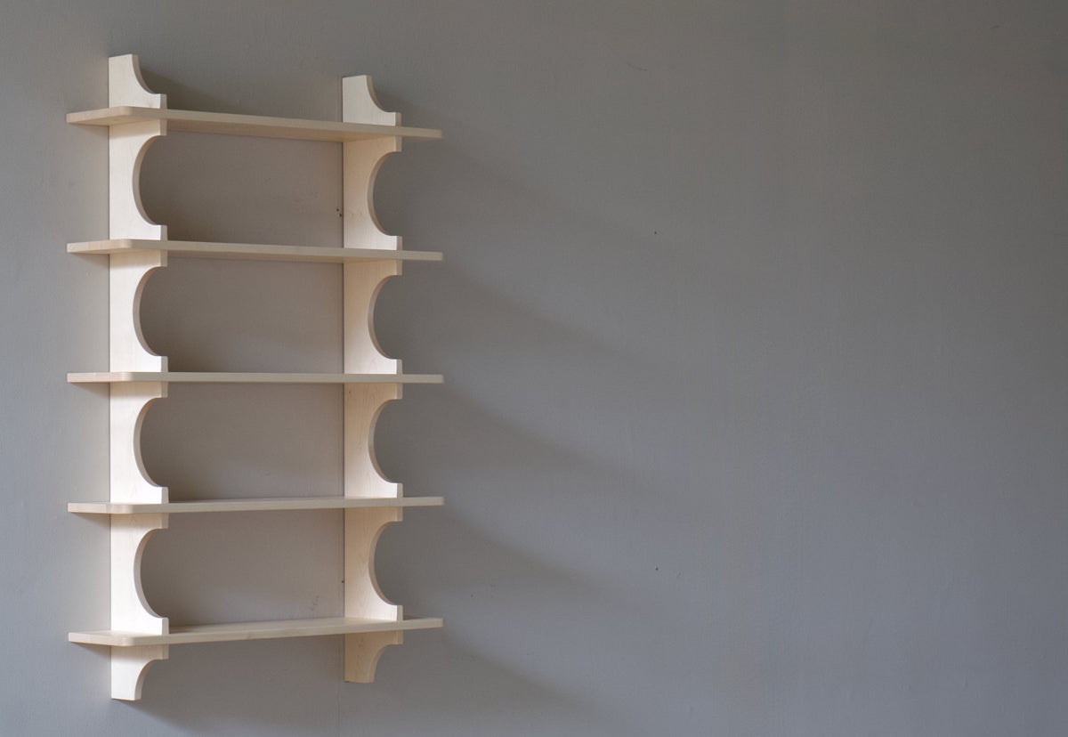 A minimalist five-tier wooden wall shelf with curved cutout details, mounted on a plain gray wall above a rustic wood floor.