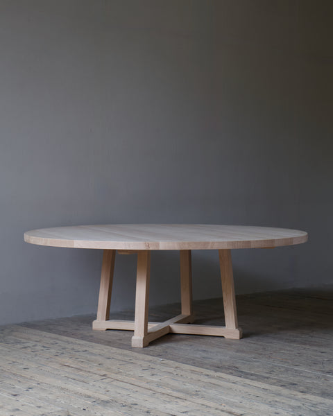 A circular light oak dining table with a minimalist trestle base, set against a dark grey wall and rustic wooden floor.