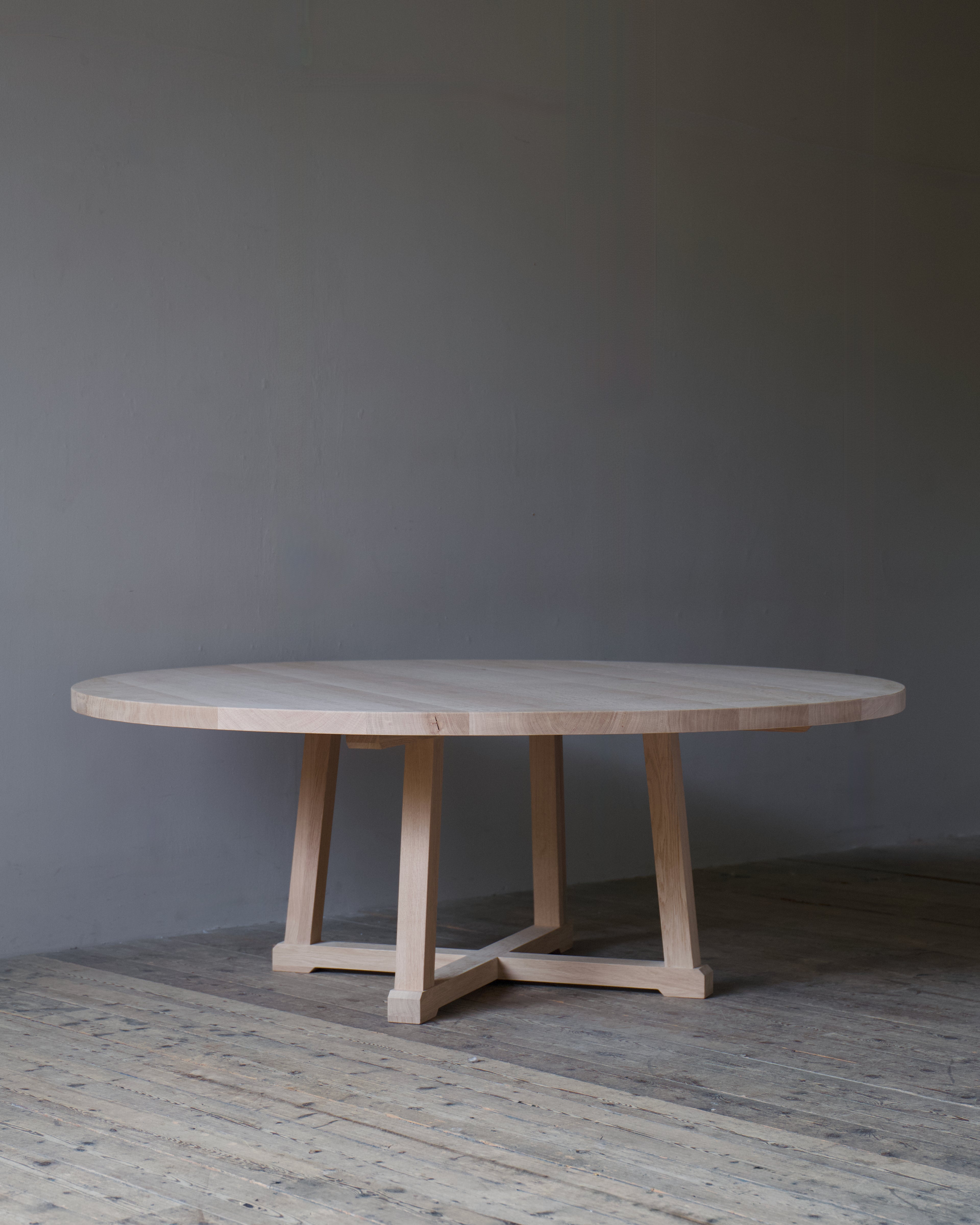 A circular light oak dining table with a minimalist trestle base, set against a dark grey wall and rustic wooden floor.