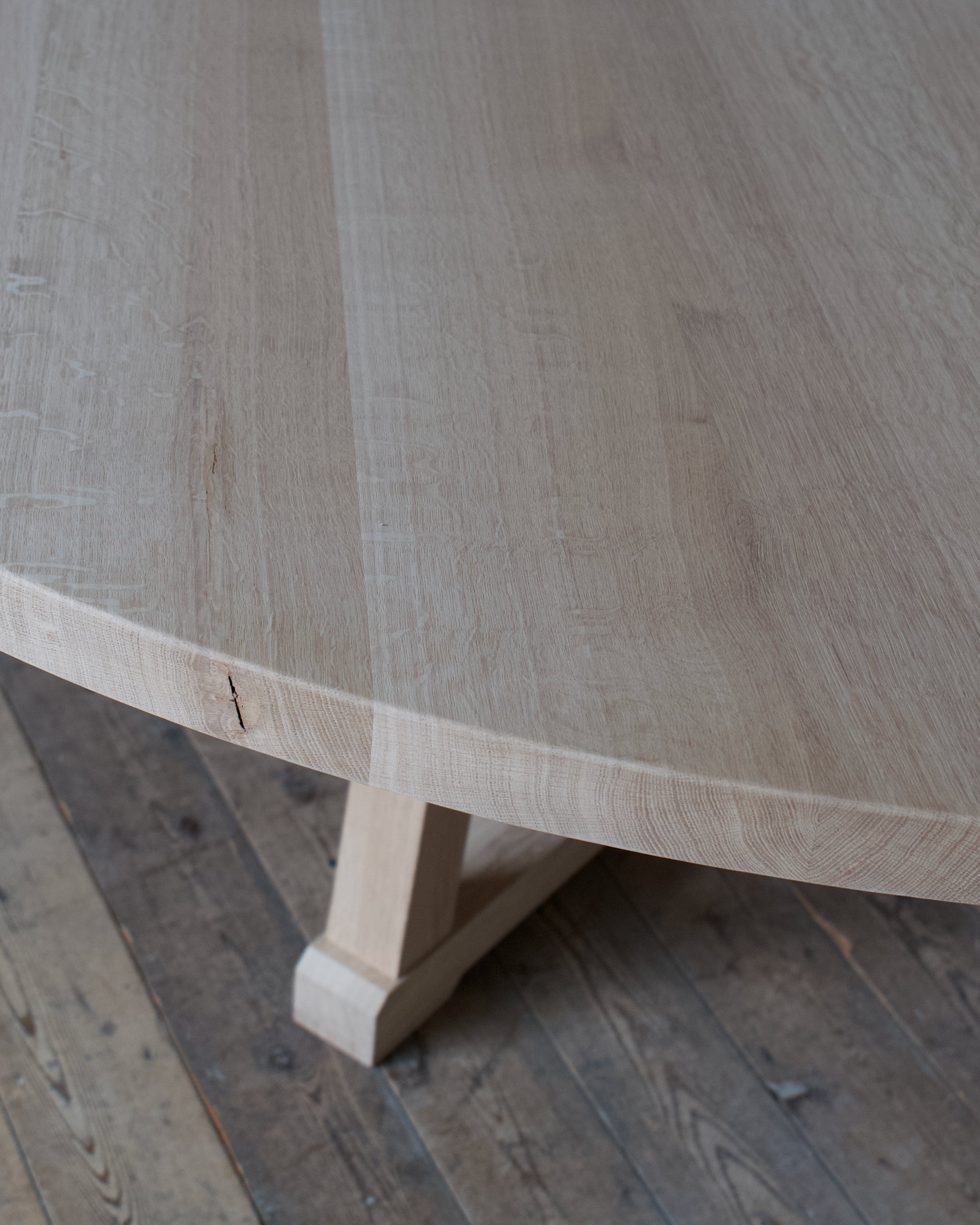 Close-up of a light oak wood dining table showing the grain and a trestle leg, set on a rustic dark wood plank floor.
