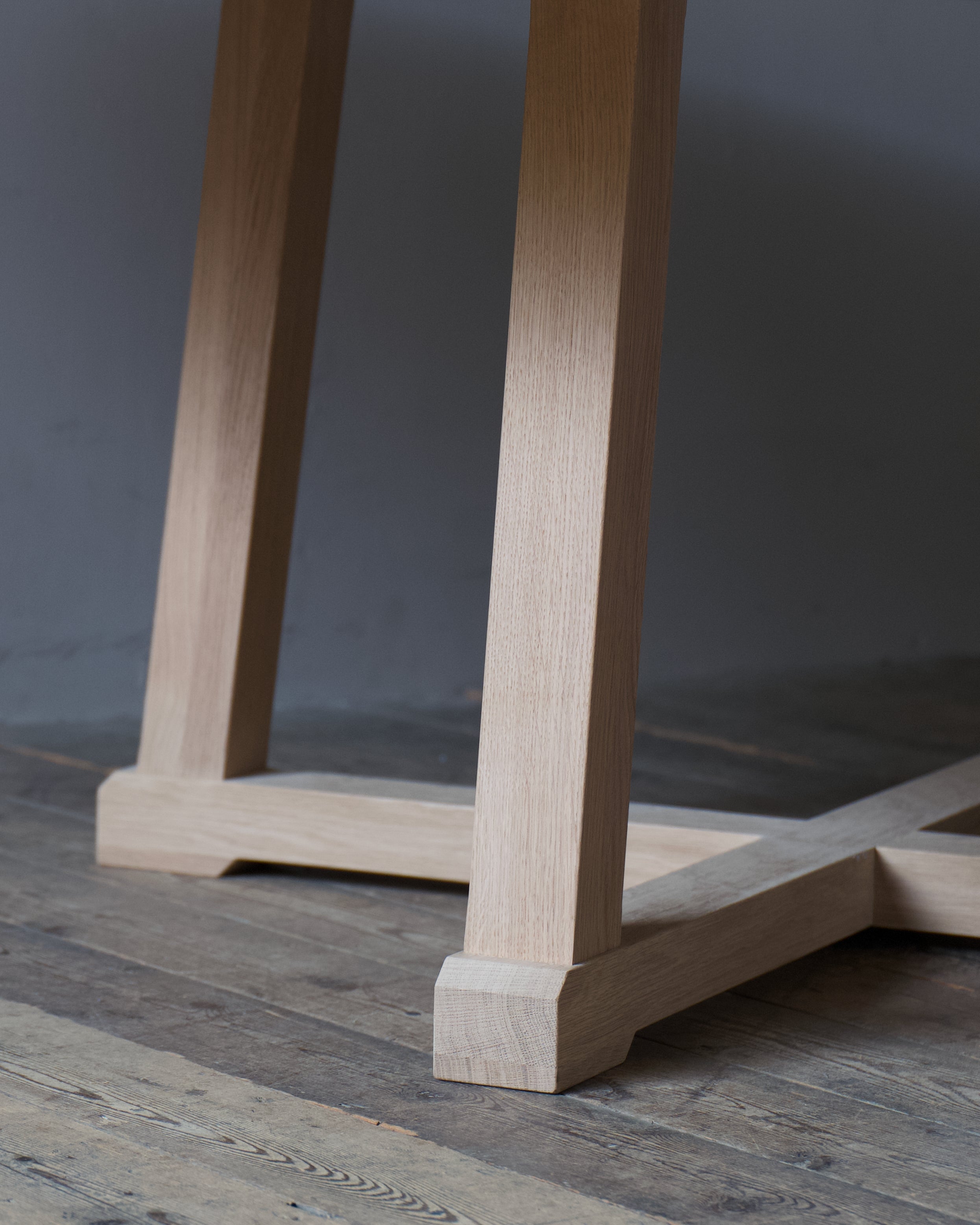 Close-up of a light oak table leg and base on a rustic dark wood floor against a dark grey wall.