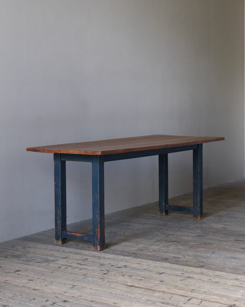 A rustic rectangular table with a medium brown wooden top and dark blue distressed legs, set against a plain gray wall and light wood floor.
