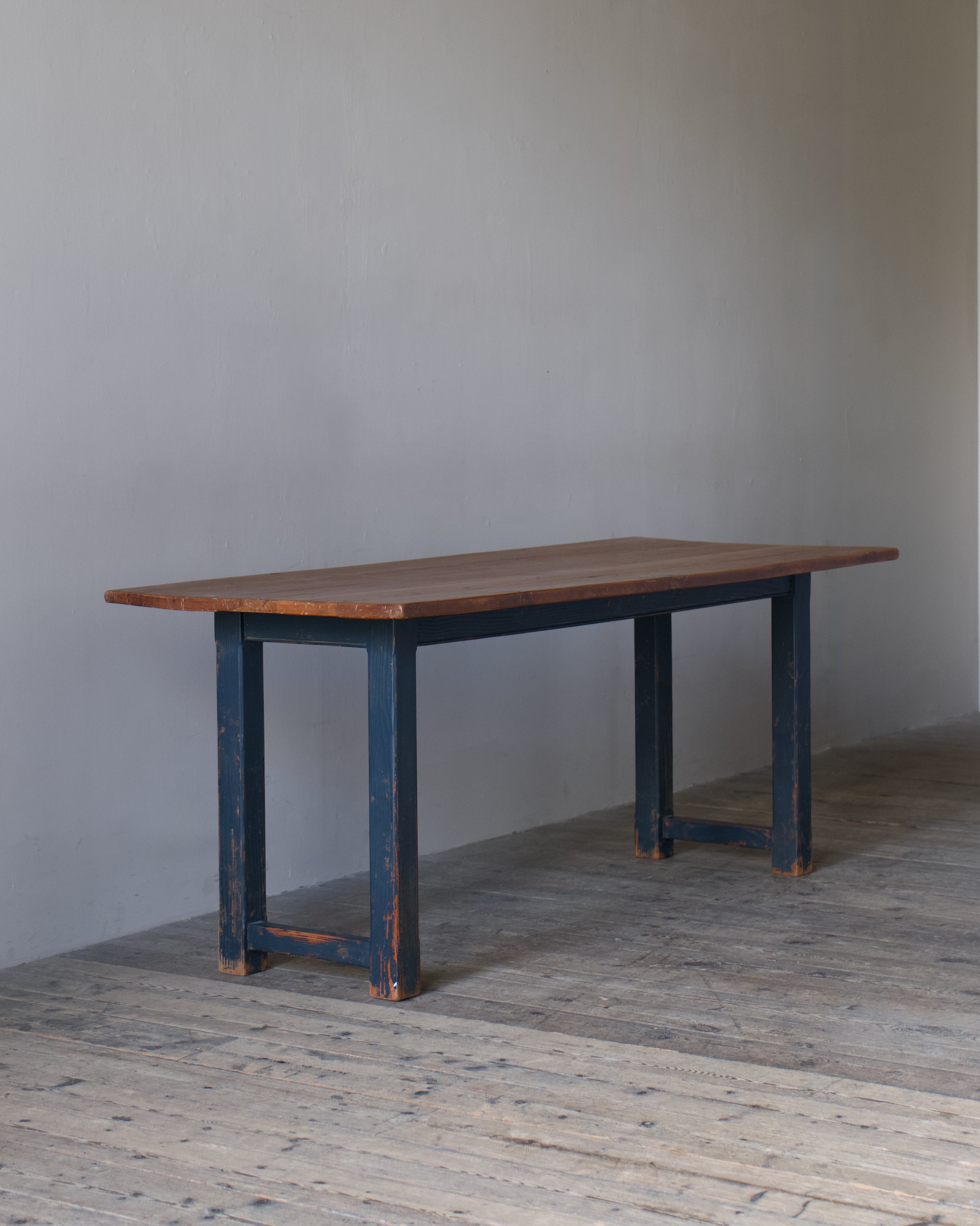 A rustic rectangular table with a medium brown wooden top and dark blue distressed legs, set against a plain gray wall and light wood floor.
