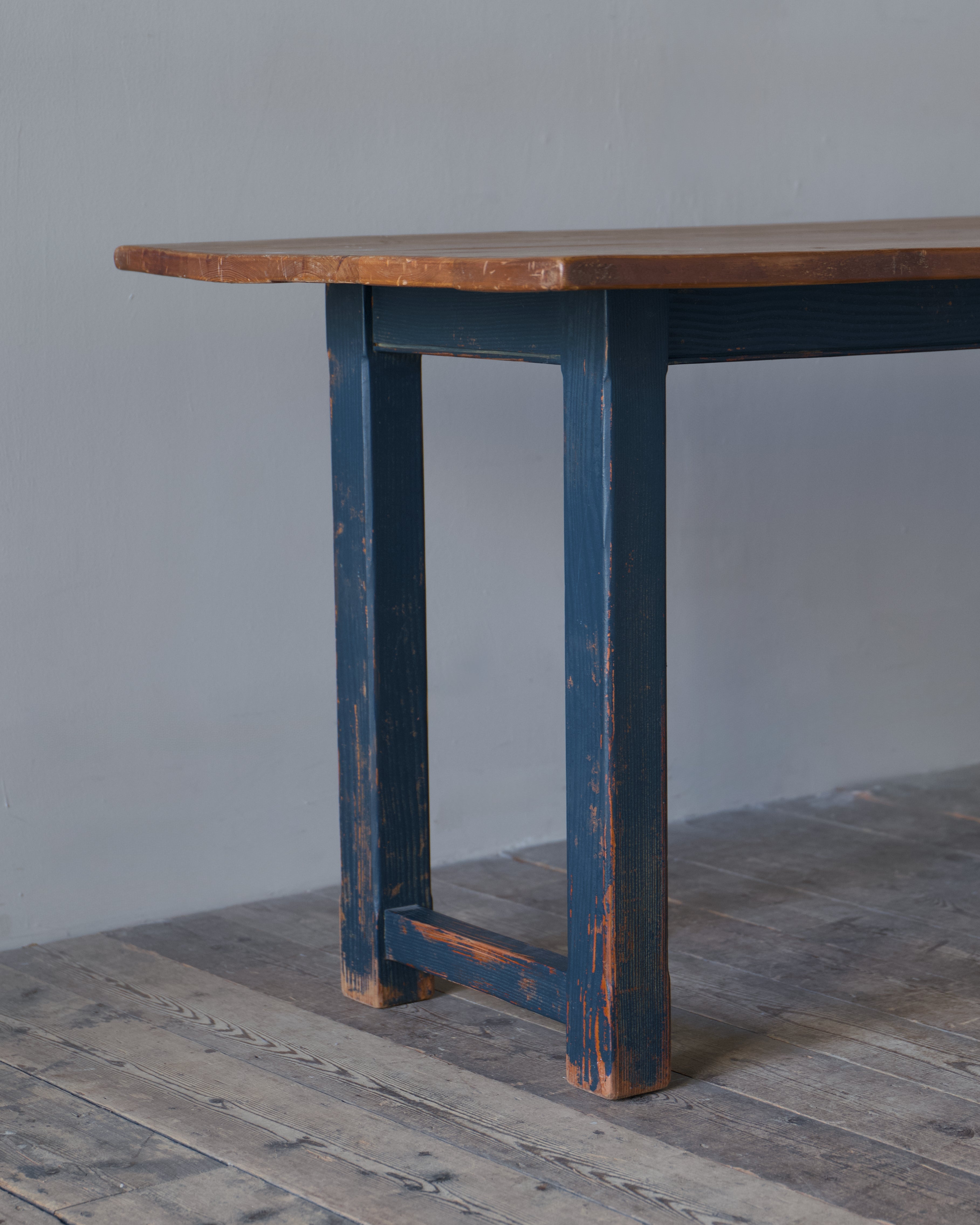 A rustic wooden table with a dark blue distressed painted frame and a natural brown wood top, shown in a minimalist room setting with a gray wall and weathered wooden floor.