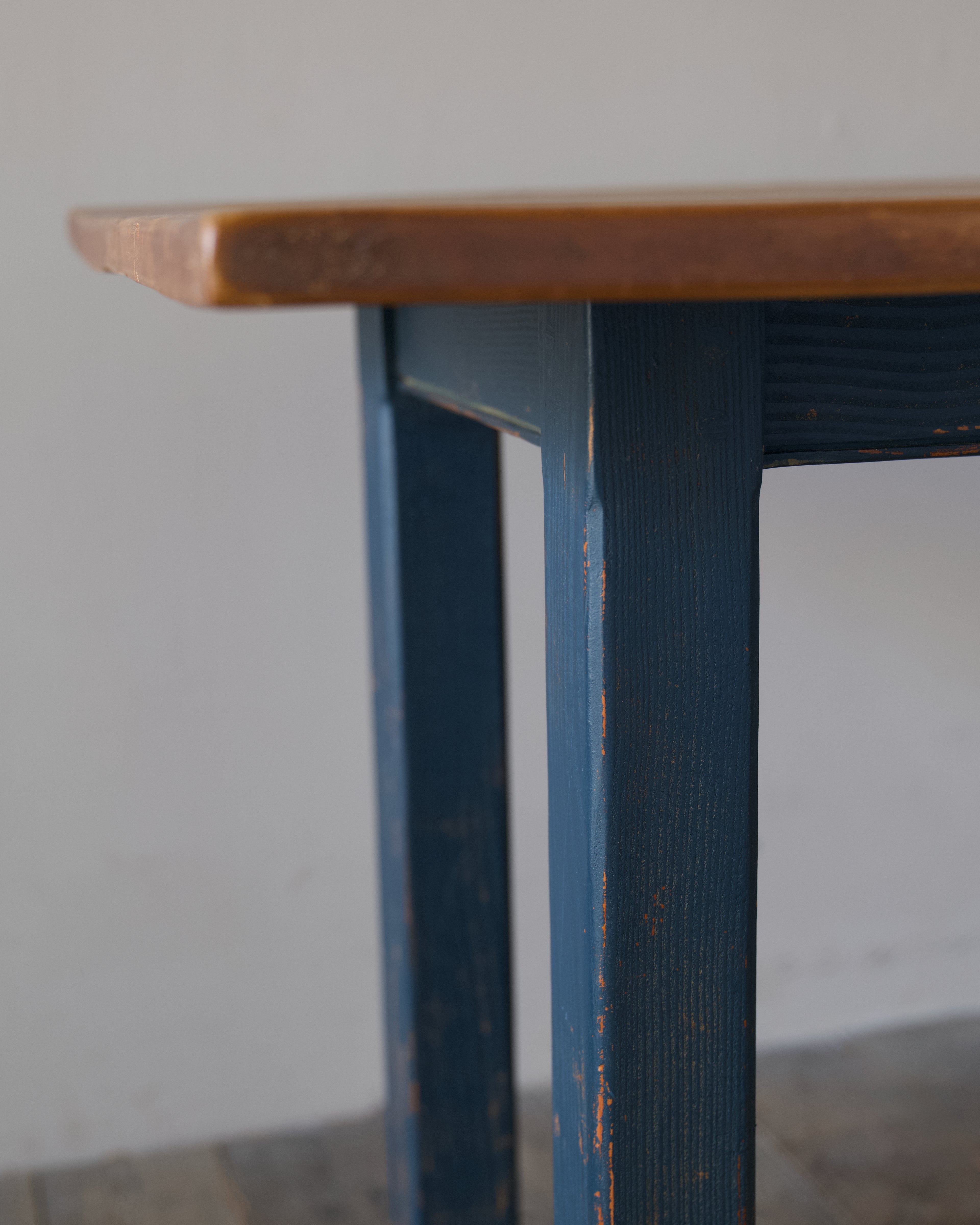 Close-up of a rustic wooden table corner with a blue distressed painted leg and a warm brown wood top against a plain light gray wall.