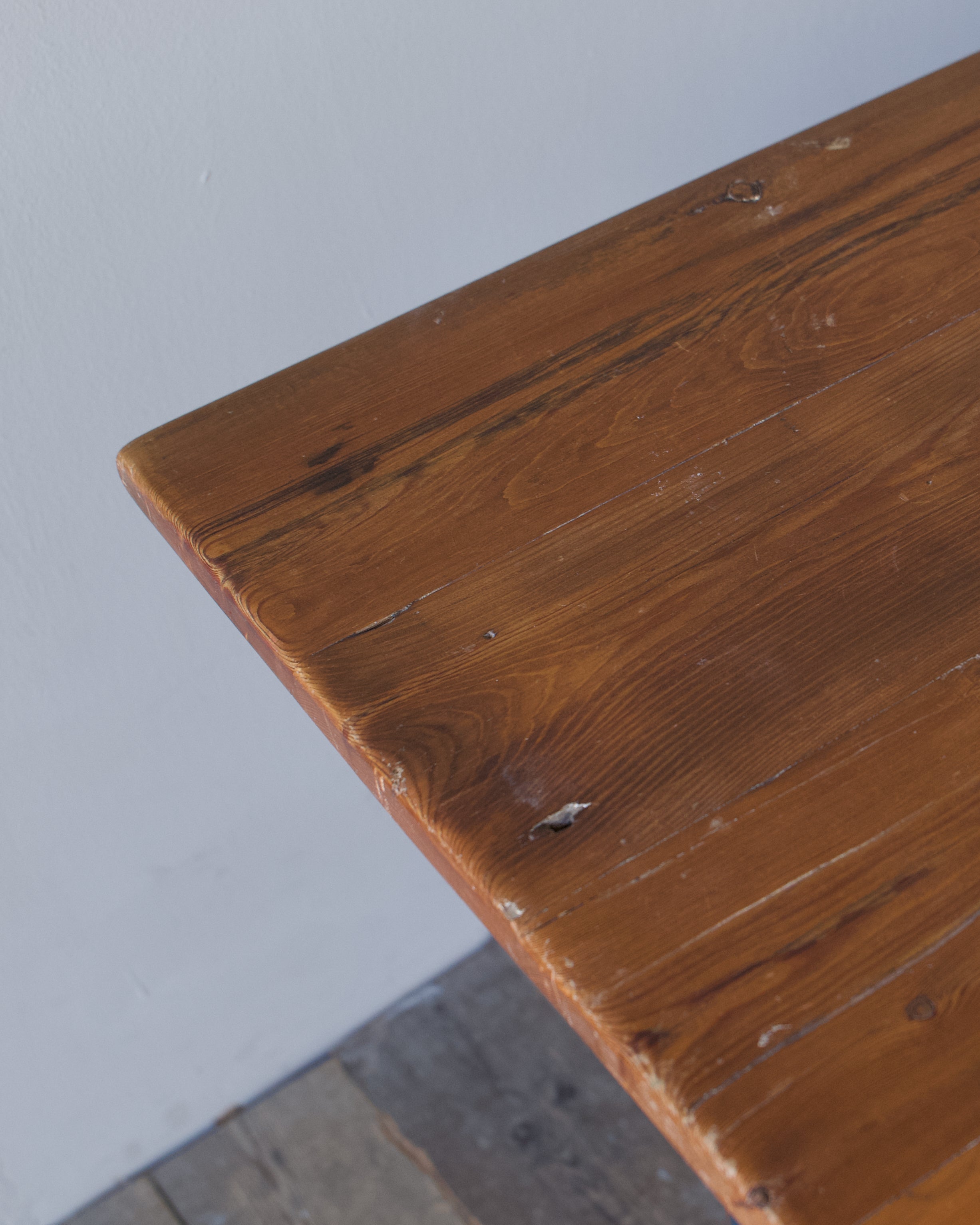 Close-up of a rustic wooden table corner showing the warm brown wood grain, knots, and weathered surface against a light gray wall and floor.