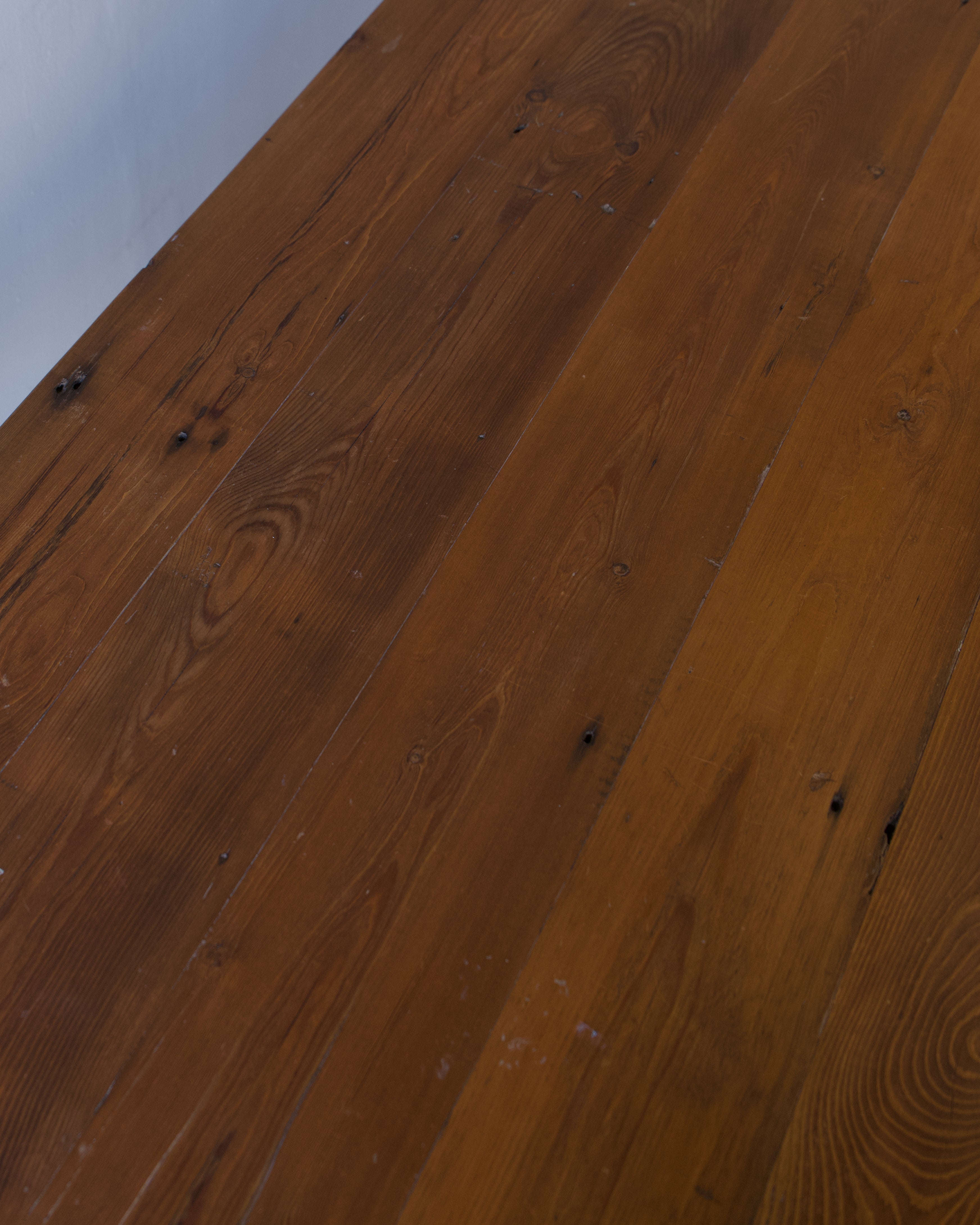 Warm medium-brown hardwood flooring planks with visible natural grain, knots, and slight wear marks, photographed at an angle against a light neutral wall.
