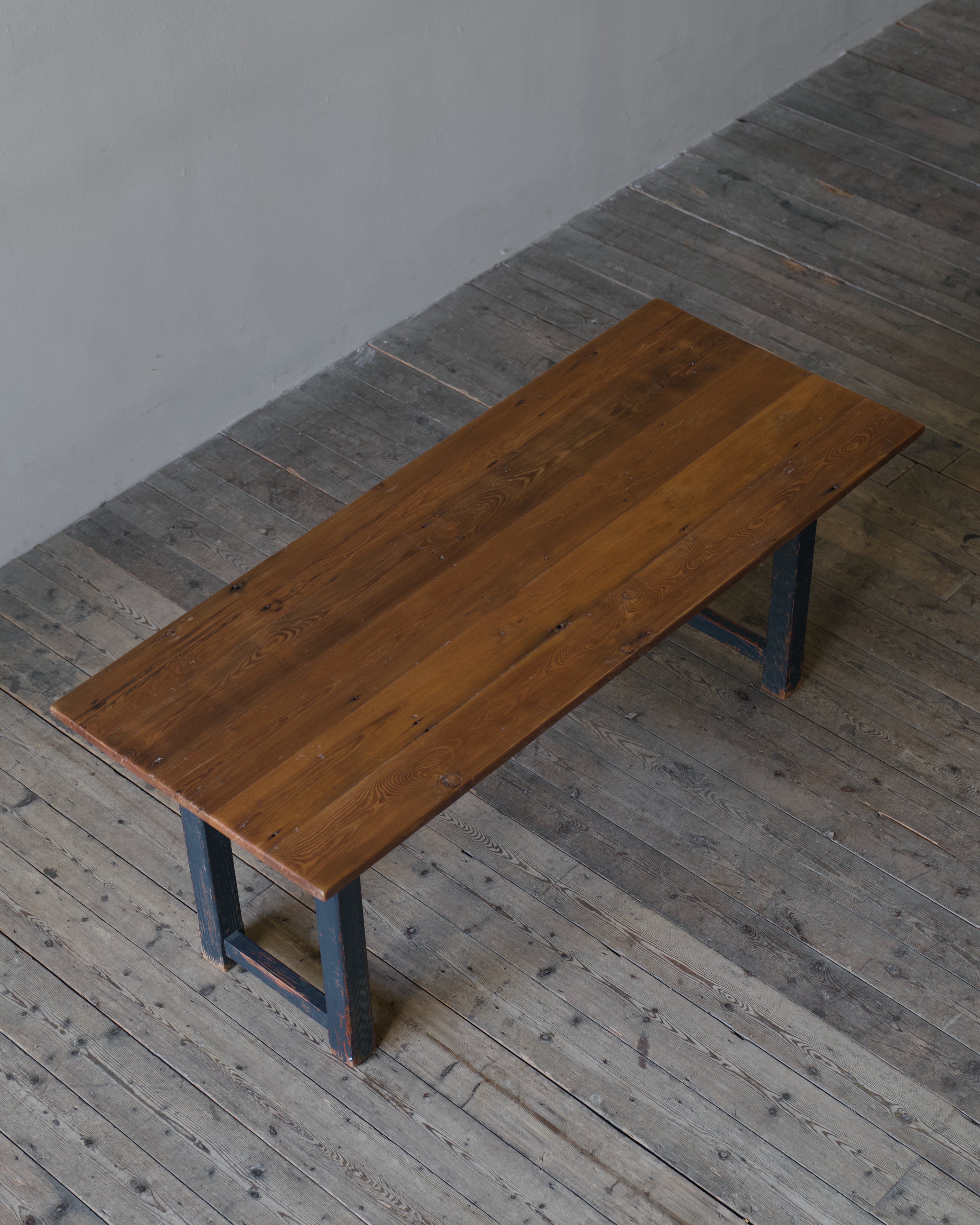 A rectangular wooden table with a warm medium-brown finish and dark blue legs, photographed in a minimalist room with grey wood floors and a light gray wall.