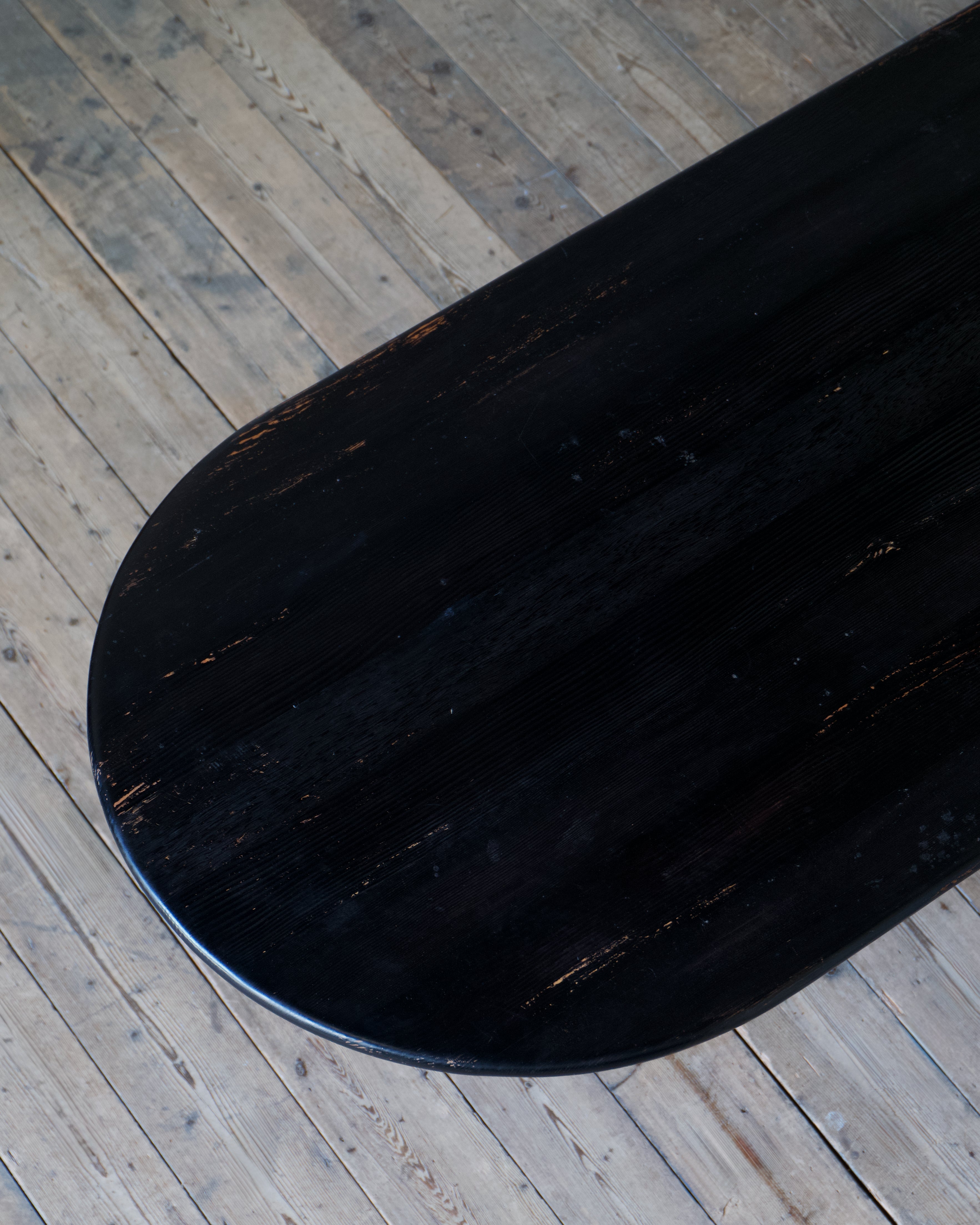 A close-up of a dark, distressed wooden board, likely a skateboard deck or table top, resting on a light-colored wooden plank floor.