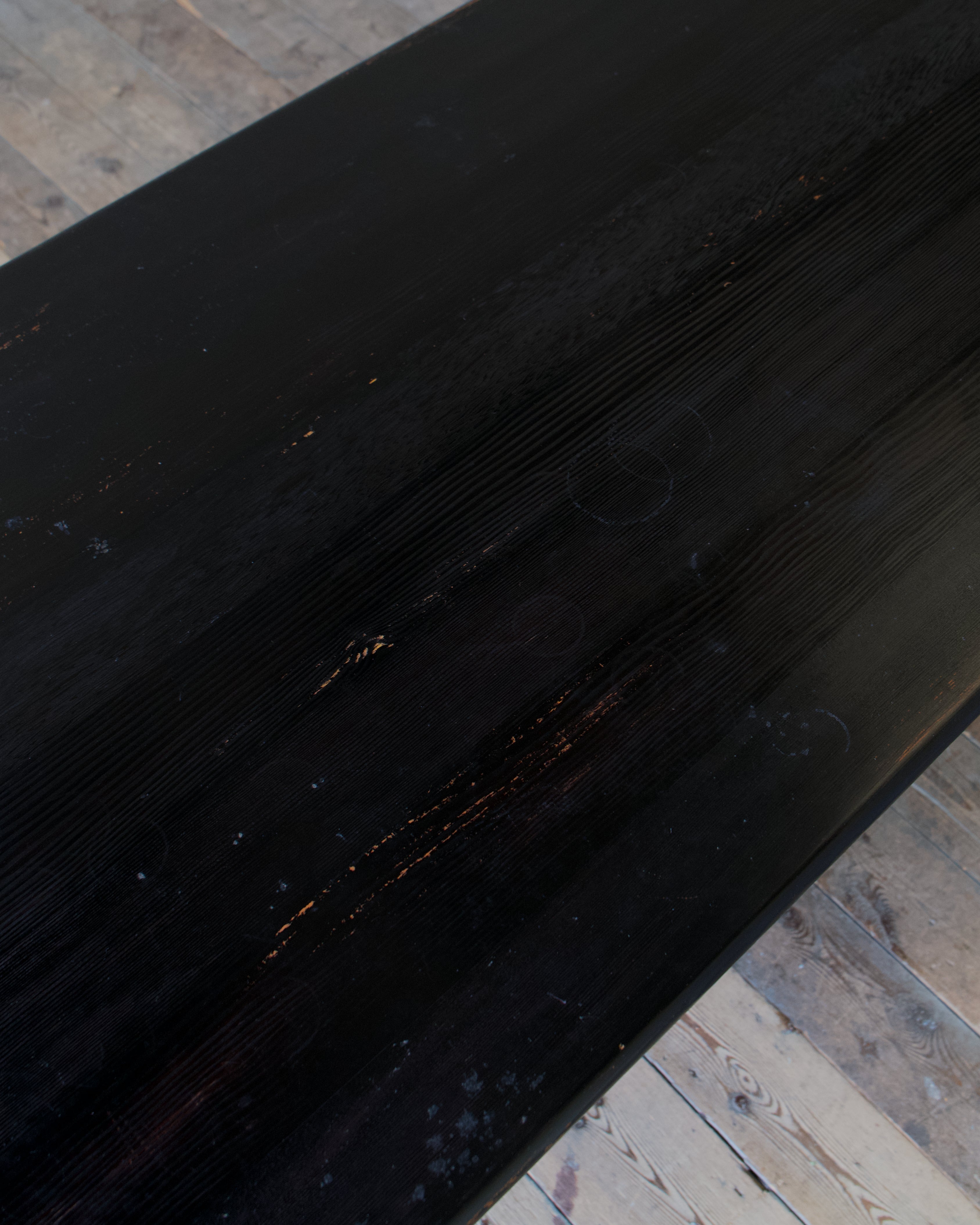 A close-up of a dark, distressed wooden surface with visible grain, scratches, and weathered texture, resting on a light wood plank floor.