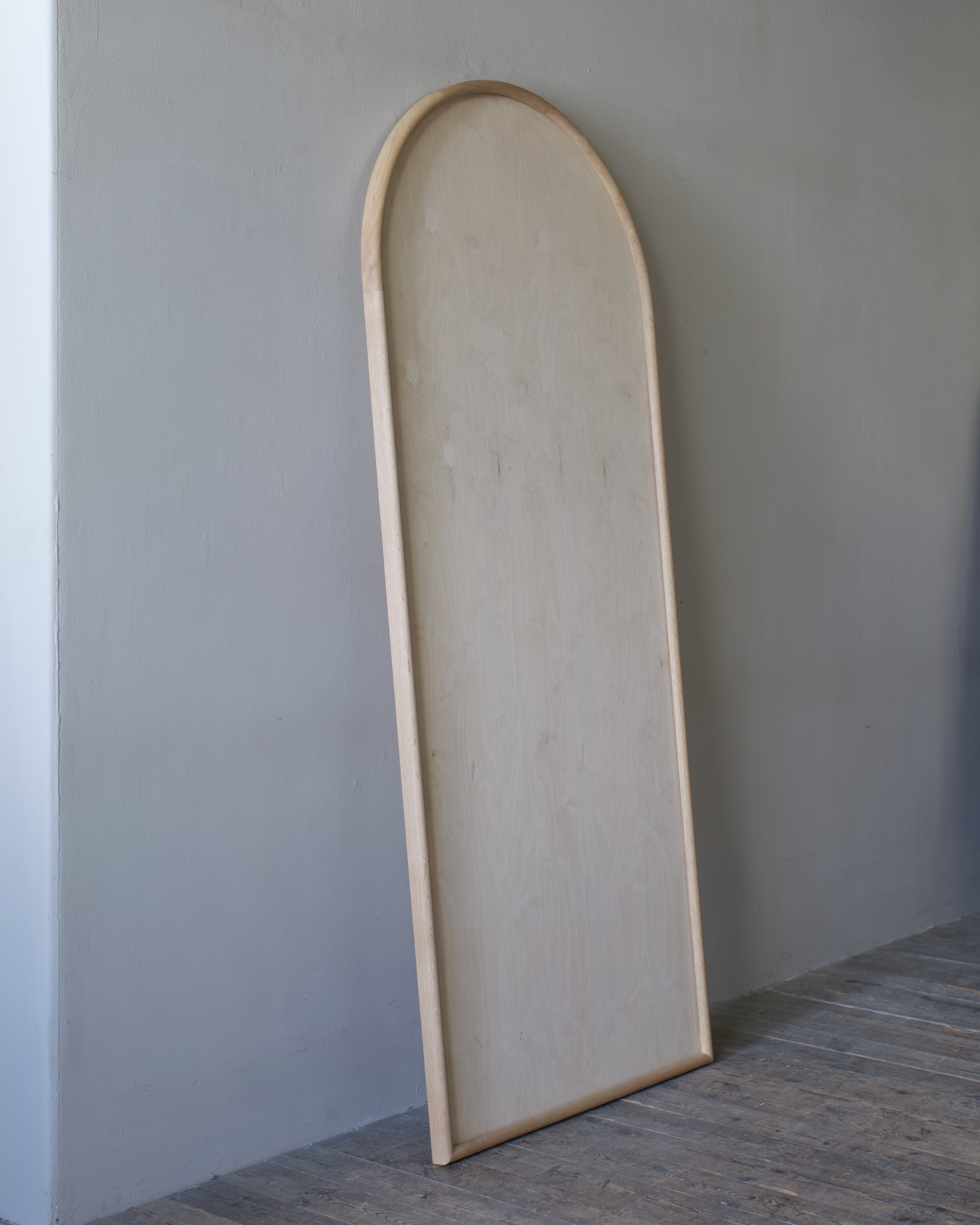 A tall, arched wooden mirror frame leaning against a gray wall on a dark wood floor.