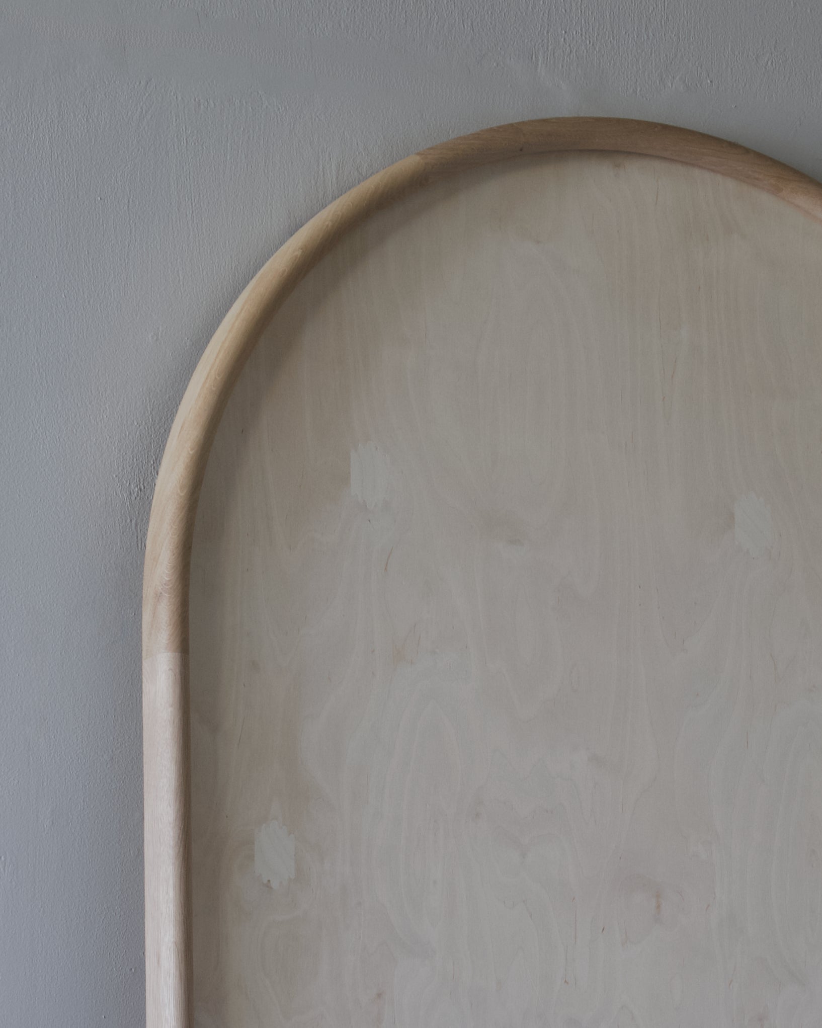 Close-up of a light natural wood arch-shaped board or tray against a textured gray wall.