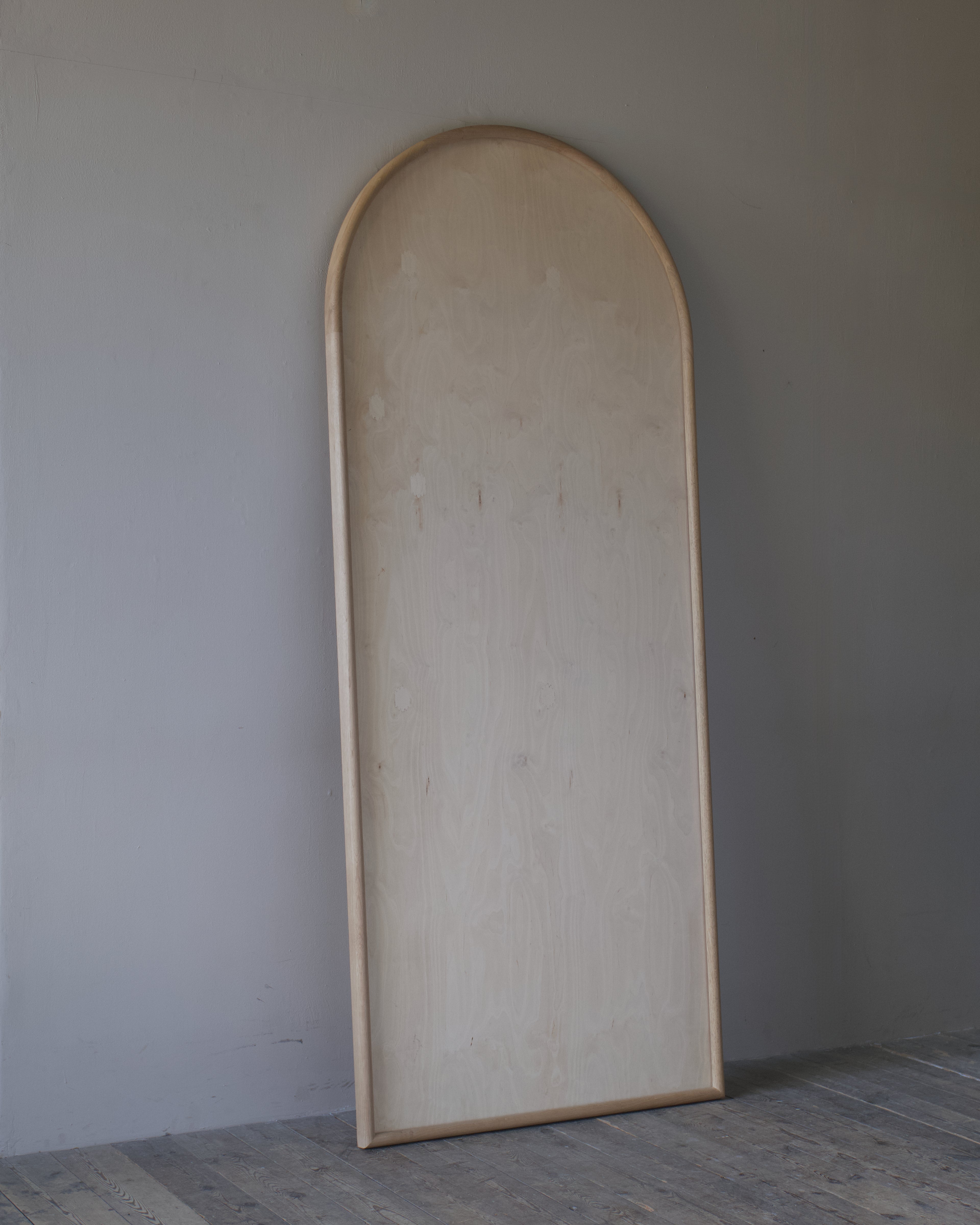 A tall, arched wooden panel or door frame made of light natural wood, leaning against a plain gray wall on a gray wooden floor.