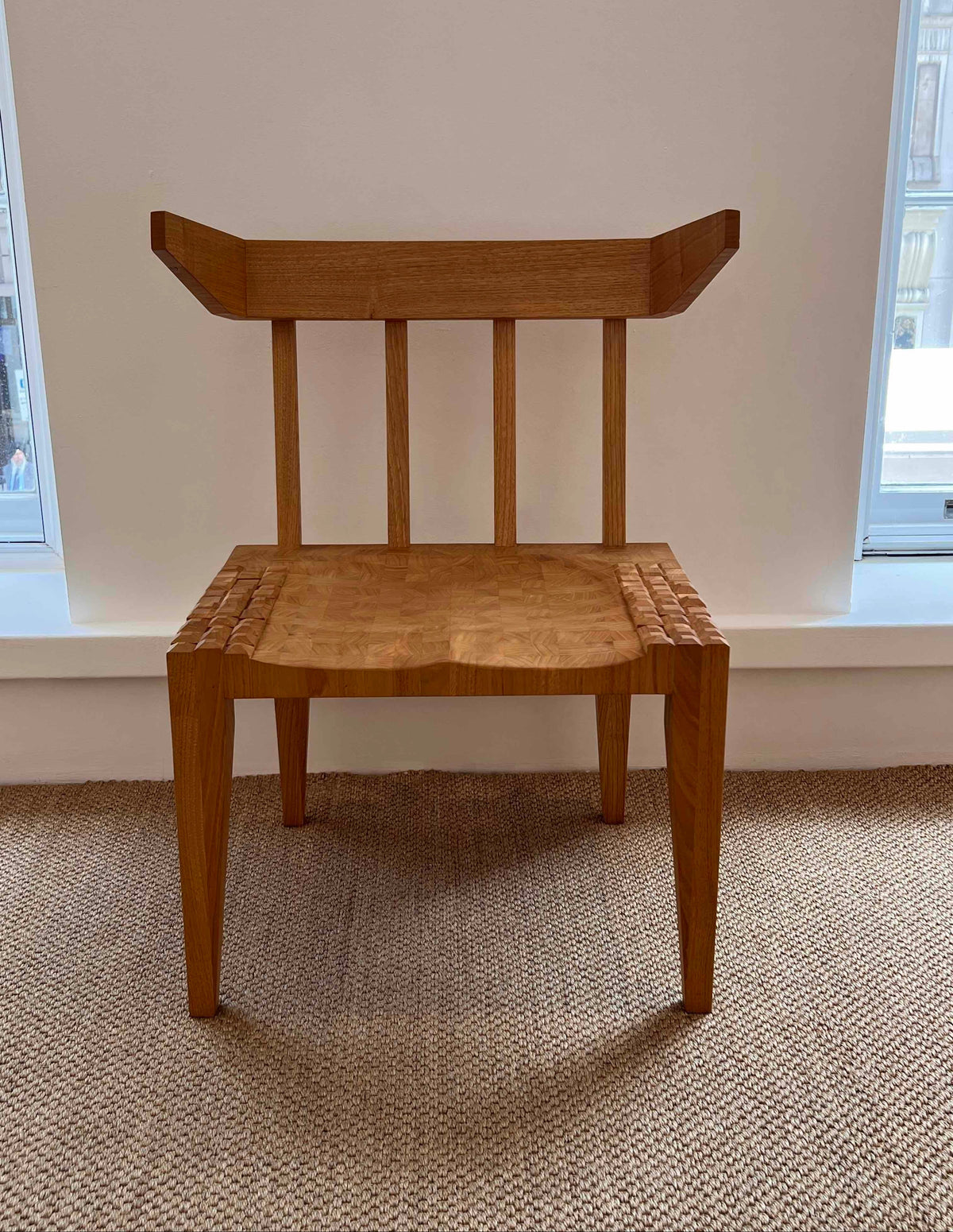 A mid-century inspired wooden dining chair with a curved backrest, vertical spindles, and a woven seat, positioned in front of a white wall with windows.