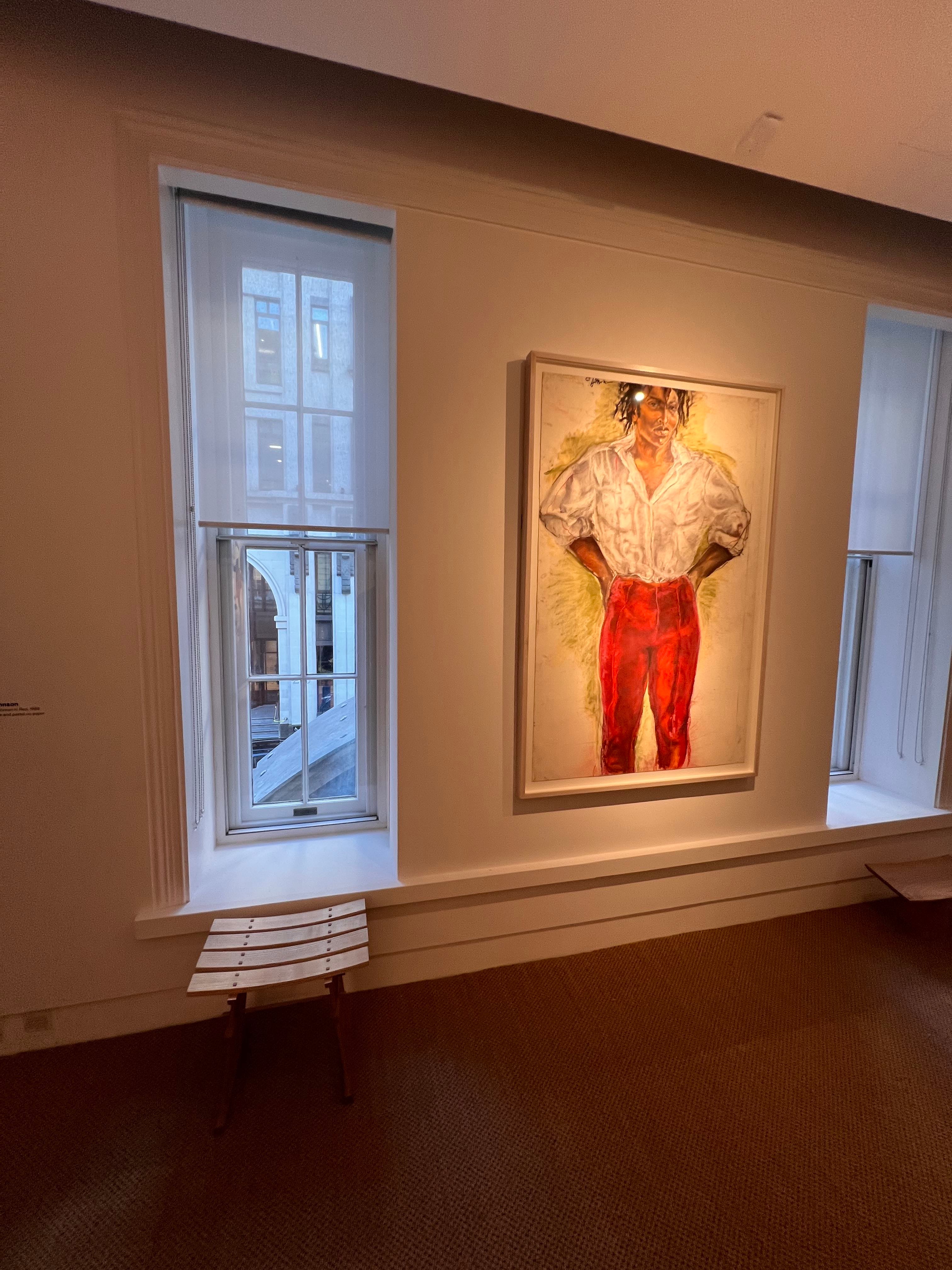 A framed portrait painting hangs on a beige gallery wall between two tall windows. The painting depicts the subject in a white shirt and bright red trousers, with warm spotlighting on the artwork.