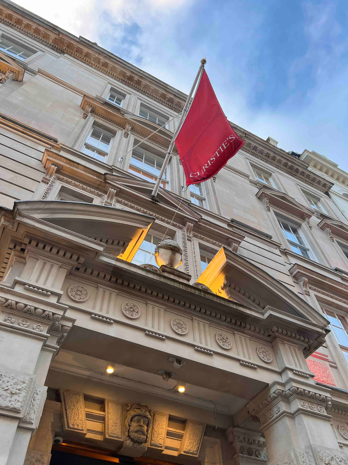 An ornate stone building facade with a large red flag reading CHRISTIES flying above a decorative entrance, set against a blue sky.