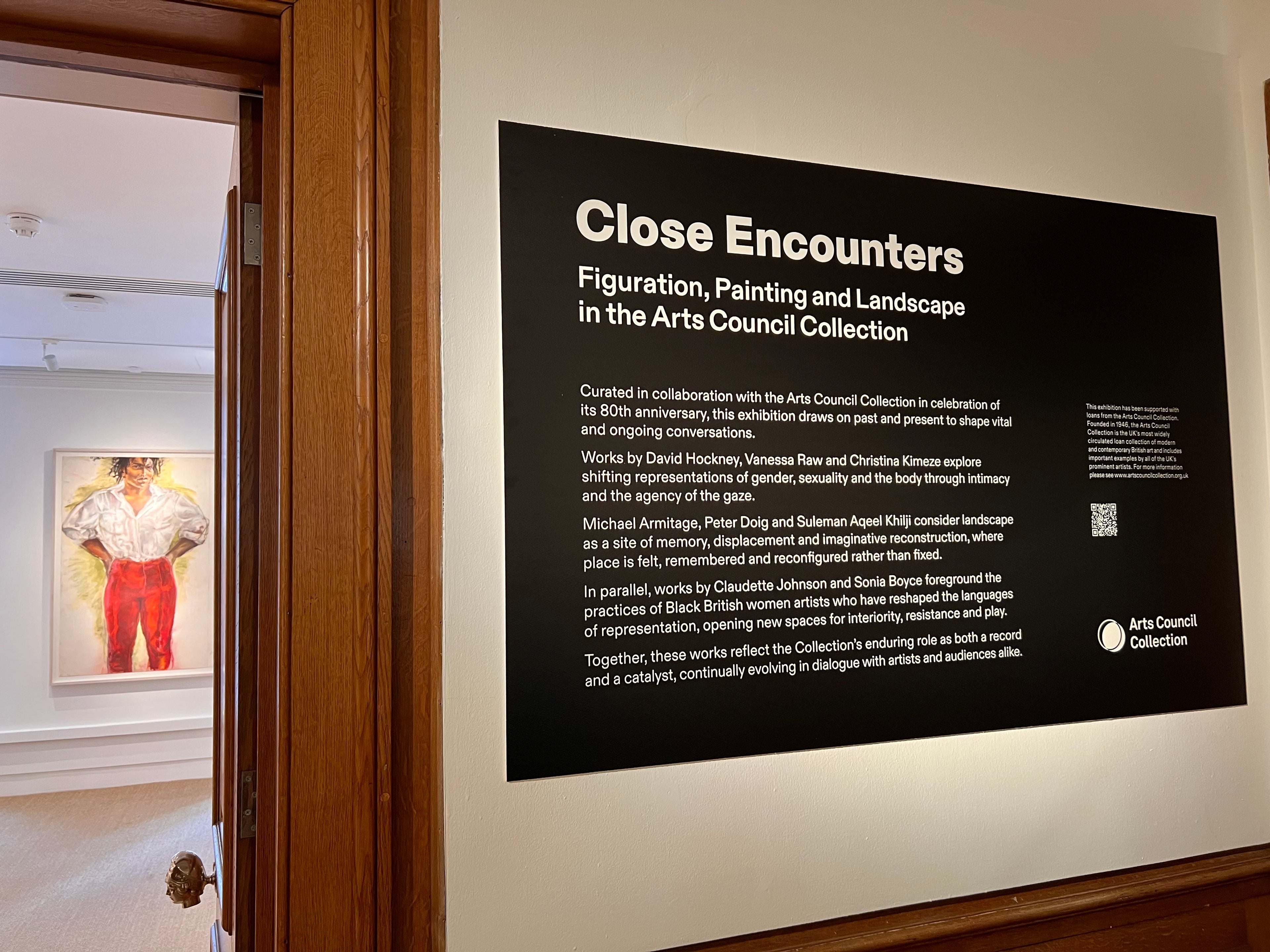 A black informational plaque with white text reading 'Close Encounters' and 'Figuration, Painting and Landscape in the Arts Council Collection,' mounted on a light wall in a real indoor environment. A doorway on the left shows part of a room with framed art.