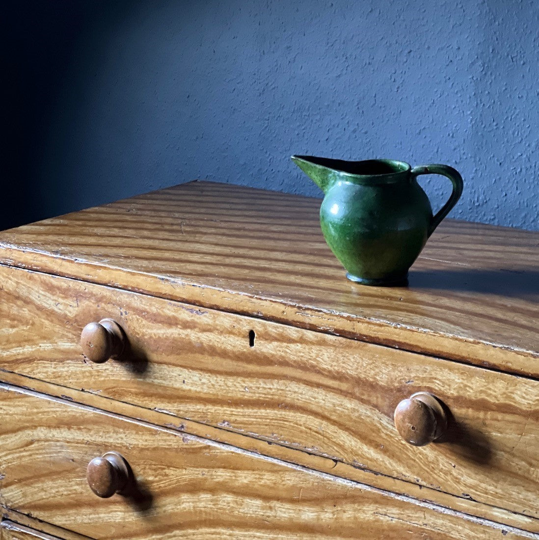 Green ceramic pitcher on a wooden chest of drawers with a blue wall background