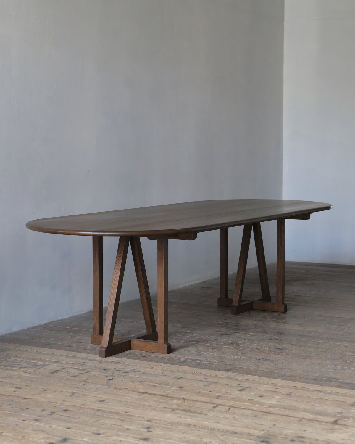 An oval oak legacy dining table with a dark fumed finish and A-frame trestle legs, set in a minimalist room with a concrete wall and wooden floor.