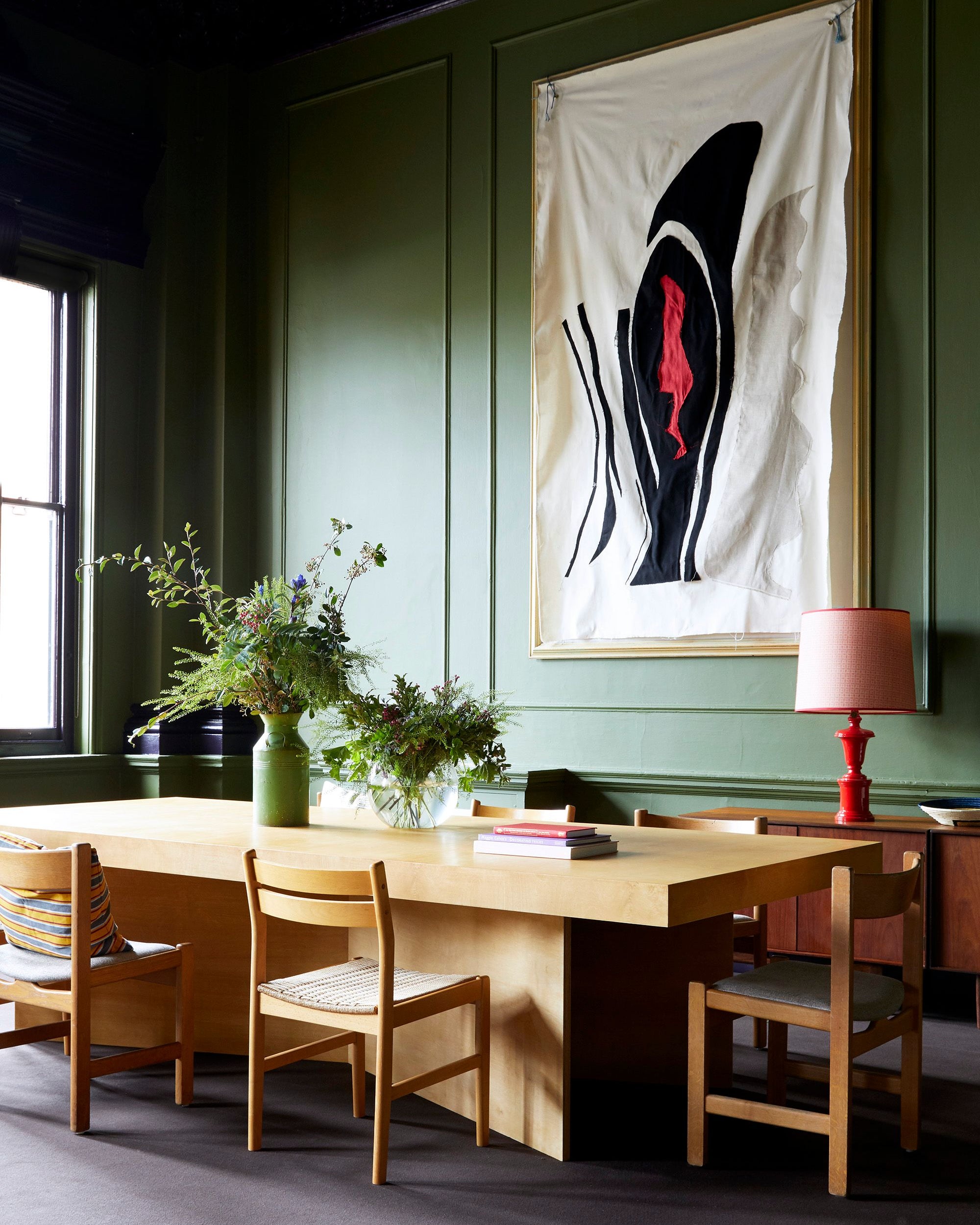 A styled interior room with dark green paneled walls, a light wood dining table, wooden chairs, a pink table lamp, and a large framed abstract wall hanging.